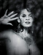 Bogini Portret zza zaparowanej szyby, jakby otulona miękką mgiełką kropel. Lekko uniesiona dłoń i spokojny wyraz twarzy tworzą atmosferę ciszy i kruchości. Całość ma eteryczny, niemal senny charakter.