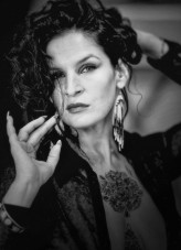 Bogini Na zdjęciu widać osobę w zbliżeniu, ukazaną w miękkim, czarno-białym tonie, który nadaje całości subtelności i elegancji. Delikatny gest dłoni, niesforne kosmyki włosów i spokojne, pewne spojrzenie tworzą atmosferę lekkości i intymne