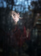 Bogini subtelne odbicie oknie, jakby zanurzone w miękkim półmroku. Delikatne światło wydobywa twarz i fragment czerwonej tkaniny, podczas gdy reszta kadru rozpływa się w refleksach i cieniach. Całość ma spokojny, intymny charakter — jak moment z